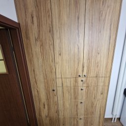 Krzysztof Mędrek - Wysoka szafa z drewnopodobnymi frontami, z trzema drzwiami na górze i trzema szufladami na dole, widoczna obok brązowych drzwi w mieszkaniu.