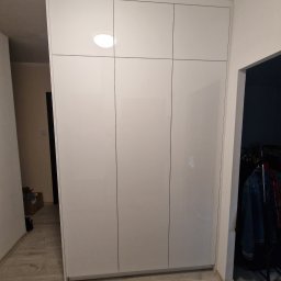 Krzysztof Mędrek - Biała, lakierowana szafa wnękowa z sześcioma drzwiami, wbudowana w przedpokoju z panelami podłogowymi w odcieniach szarości i beżu, widok z perspektywy korytarza z otwartymi drzwiami do innego...