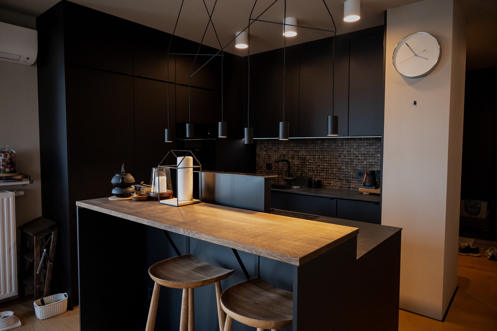 Nowoczesna kuchnia w ciemnych barwach z drewnianym blatem wyspowym i hokerami, oświetlona minimalistycznymi lampami wiszącymi.