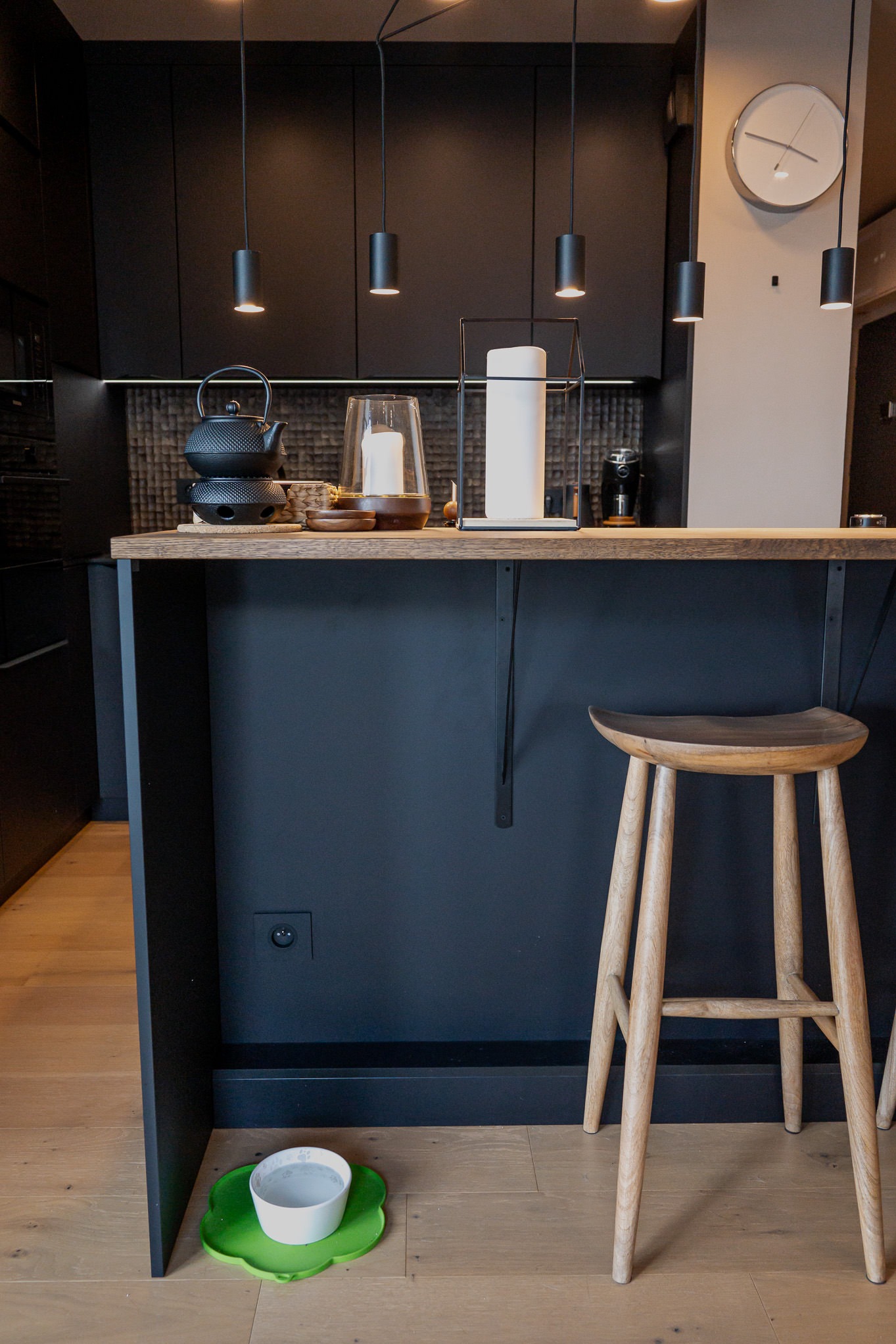 Nowoczesna kuchnia z czarnymi szafkami, drewnianym blatem i hokerami. Na blacie czajnik, świecznik i inne akcesoria. Pod blatem miska dla zwierząt na zielonej podkładce.