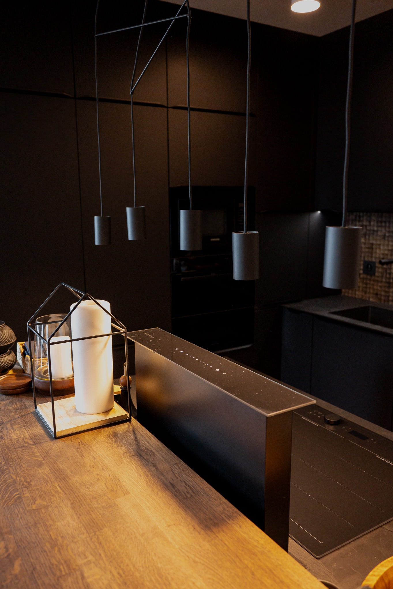 Ciemna kuchnia z drewnianym blatem, nowoczesne lampy wiszące nad blatem, dekoracyjna latarenka ze świecami, minimalistyczny design.
