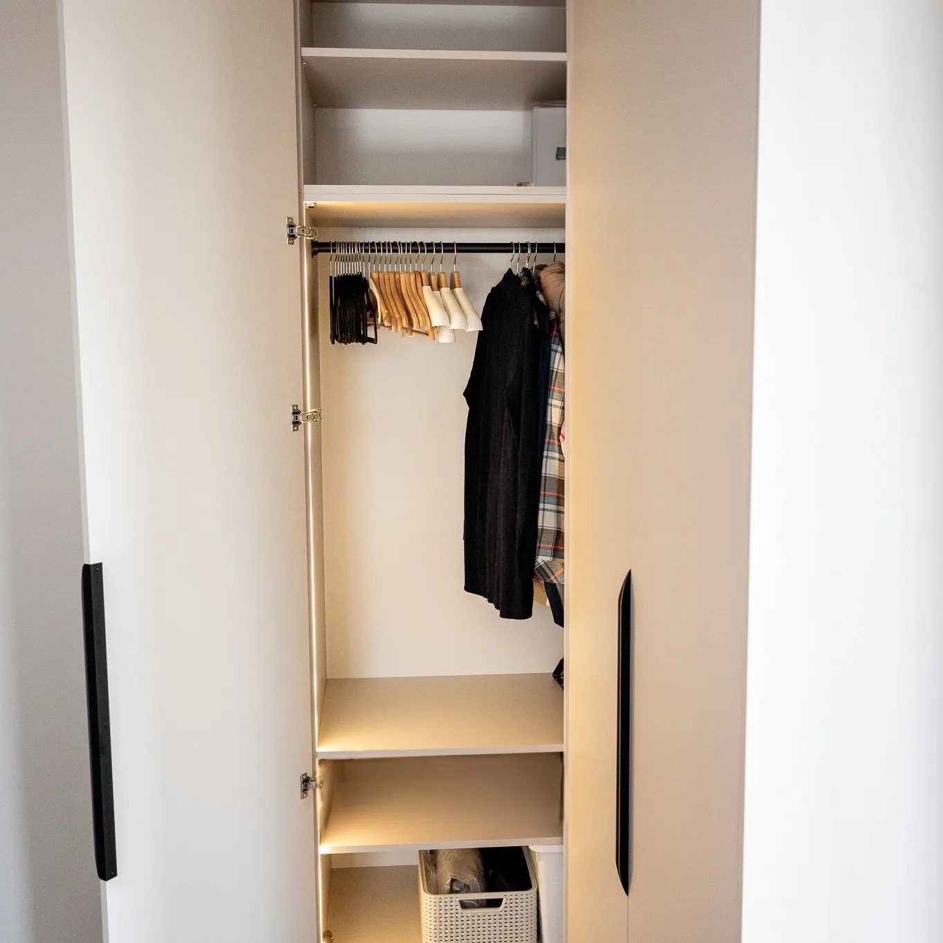Otwarta szafa wnękowa z oświetleniem LED, widoczne wieszaki z ubraniami w środku i kosz na dole, minimalistyczne czarne uchwyty.