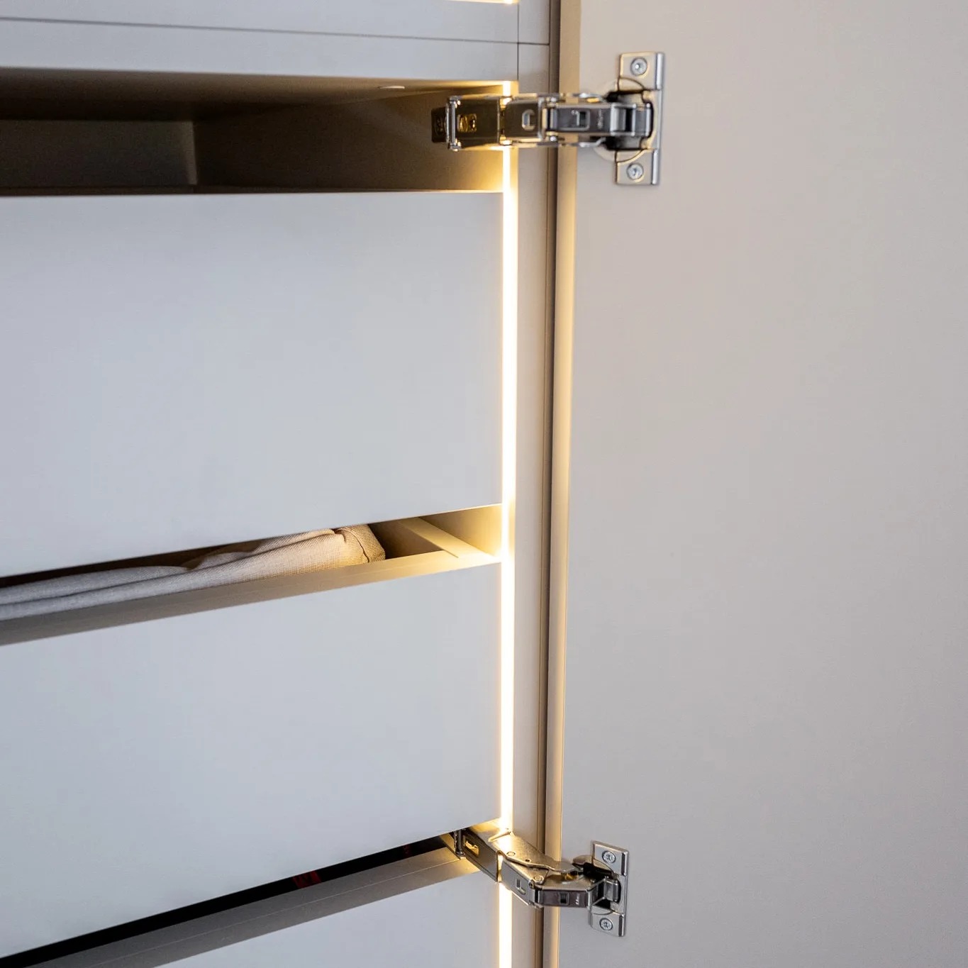 Otwarta szafa z białymi frontami szuflad, oświetleniem LED w pionowych szczelinach i widocznymi zawiasami.