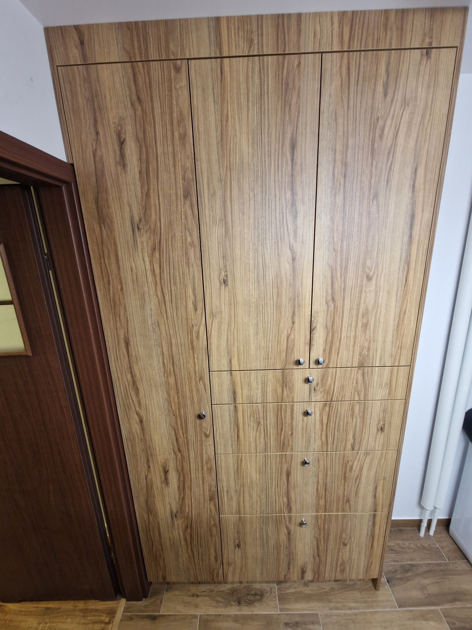 Wysoka szafa z drewnopodobnymi frontami, z trzema drzwiami na górze i trzema szufladami na dole, widoczna obok brązowych drzwi w mieszkaniu.