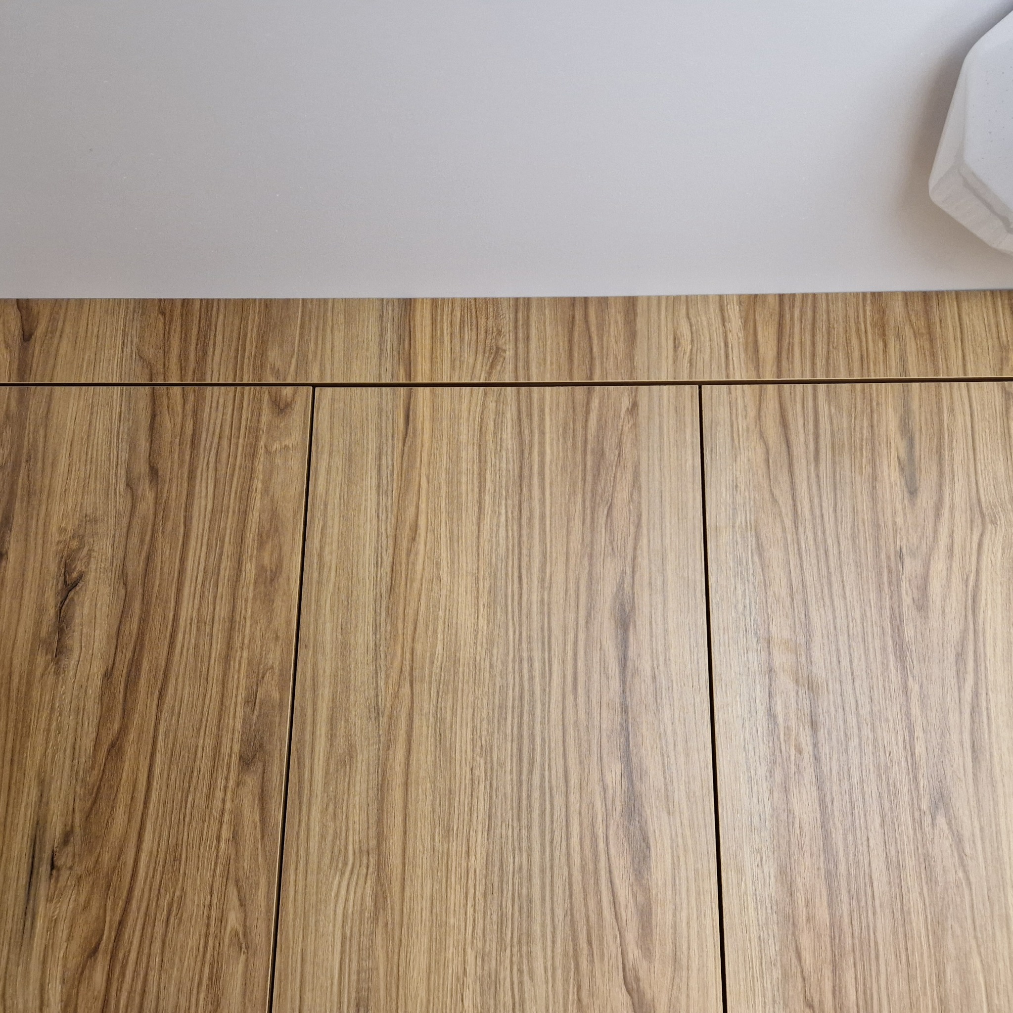 Fragment zabudowy kuchennej z drewnianymi frontami, widoczne trzy pionowe drzwi szafek z wyraźnym usłojeniem drewna, u góry biały sufit z fragmentem lampy.