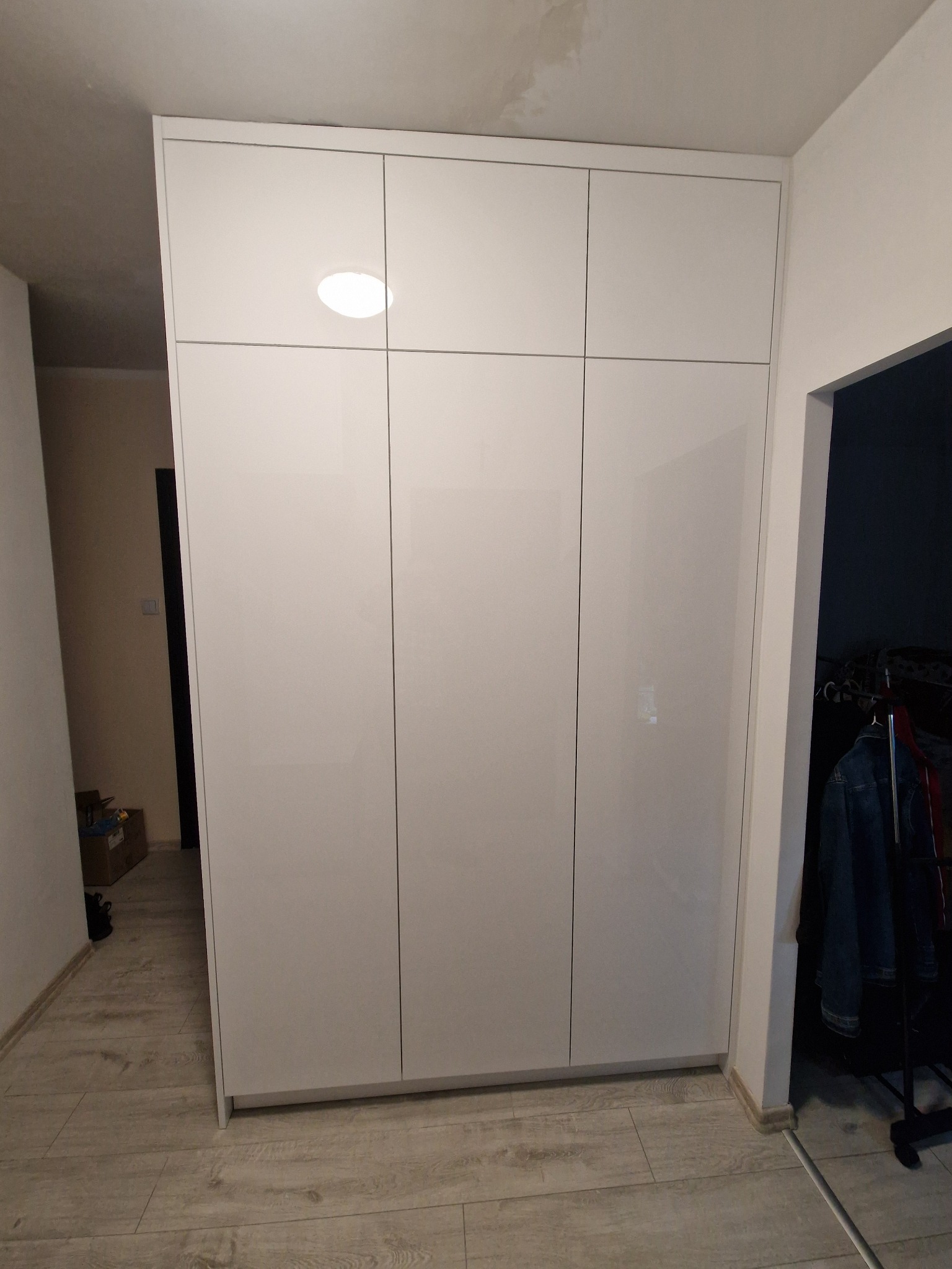 Biała, lakierowana szafa wnękowa z sześcioma drzwiami, wbudowana w przedpokoju z panelami podłogowymi w odcieniach szarości i beżu, widok z perspektywy korytarza z otwartymi drzwiami do innego...