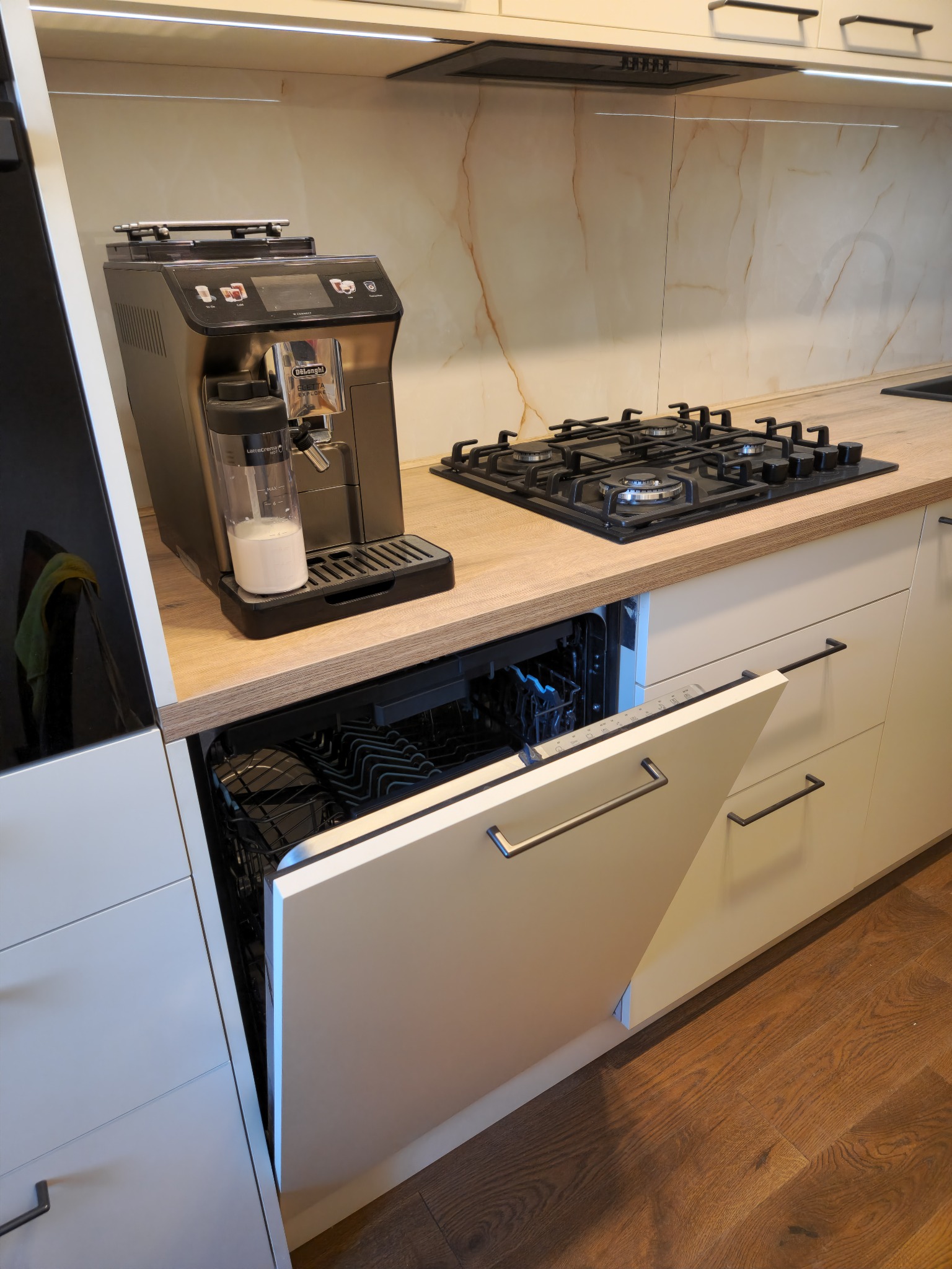 Nowoczesna kuchnia z jasnymi szafkami, drewnianym blatem, płytą gazową i otwartą zmywarką, obok ekspres do kawy z mlekiem.