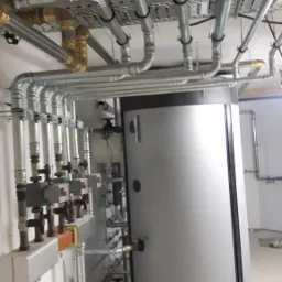 Srebrny zasobnik ciepłej wody z podłączonymi rurami miedzianymi i stalowymi, zamontowany w pomieszczeniu technicznym z widoczną instalacją pod sufitem.