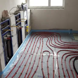 Wnętrze w trakcie prac: czerwone rury ogrzewania podłogowego ułożone na folii z rastrem, widoczne rozdzielacze na ścianie, surowy stan wykończenia.