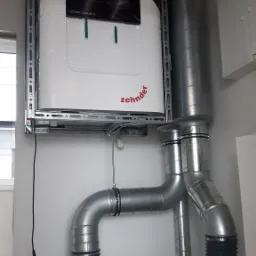 Centrala wentylacyjna Zehnder ComfoAir Q zamontowana na ścianie, z widocznymi przewodami wentylacyjnymi i elementami montażowymi, jeszcze w folii ochronnej.