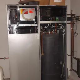 Otwarty piec dwufunkcyjny z widocznym zasobnikiem wody, instalacja z rur miedzianych na ścianie i suficie, kartony na górze pieca, kanister z płynem obok, widok z bliska.