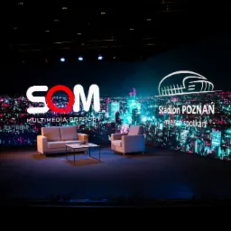 Scena z ekranem LED wyświetlającym panoramę miasta nocą, logo 'SOM MULTIMEDIA SUPPORT' i 'Stadion Poznań miejsce spotkań', ustawiona z beżową sofą, fotelem i stolikiem kawowym.