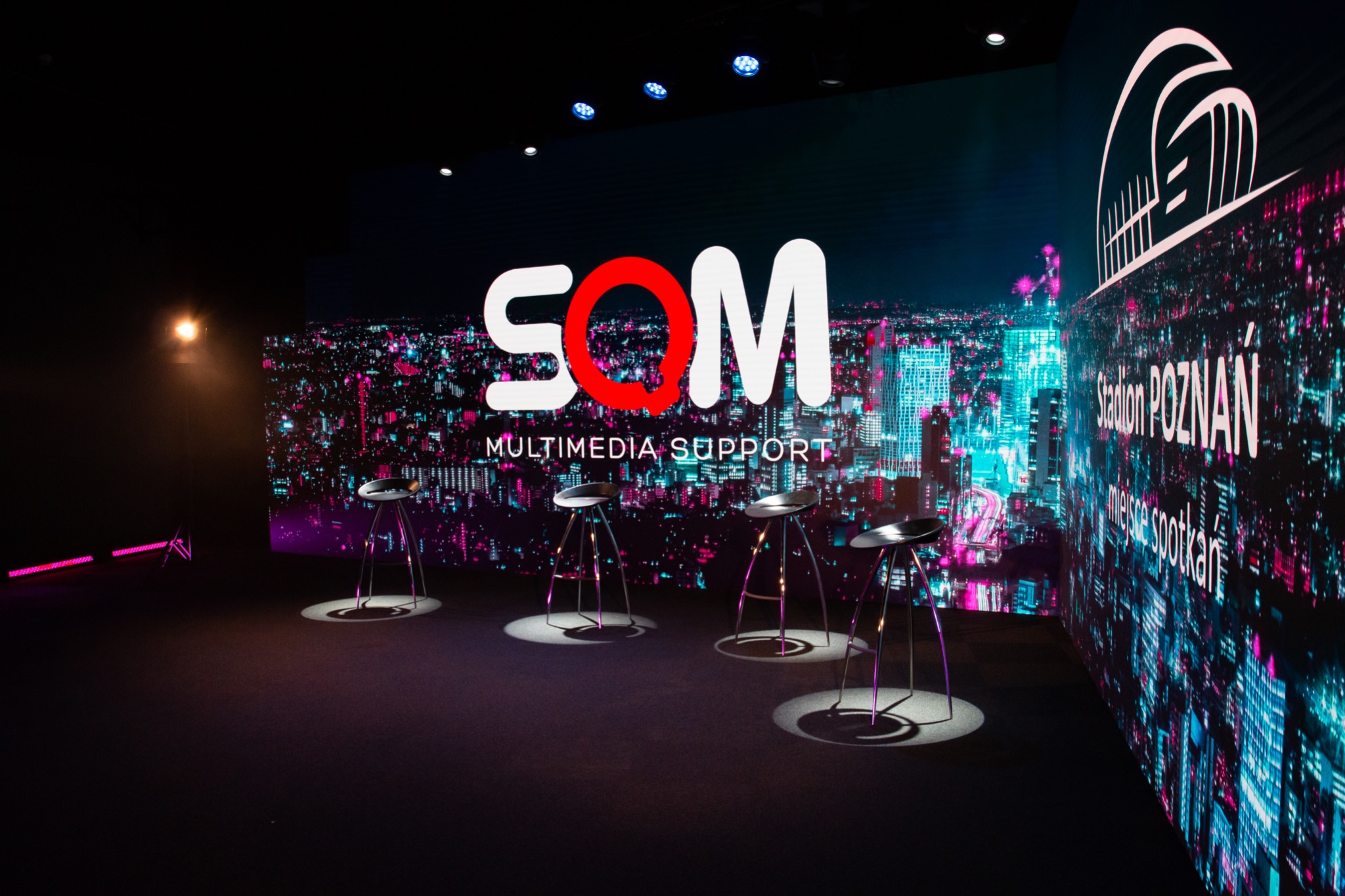Sala konferencyjna z krzesłami barowymi na tle wyświetlanych grafik miejskich i logo SOM Multimedia Support, oświetlenie punktowe na podłodze.