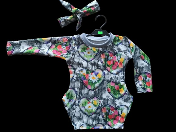 Dziecięce body z długim rękawem, w szaro-białe tło i wzór serc z tulipanami i stokrotkami, zawieszone na czarnym wieszaku, z pasującą opaską na głowę.