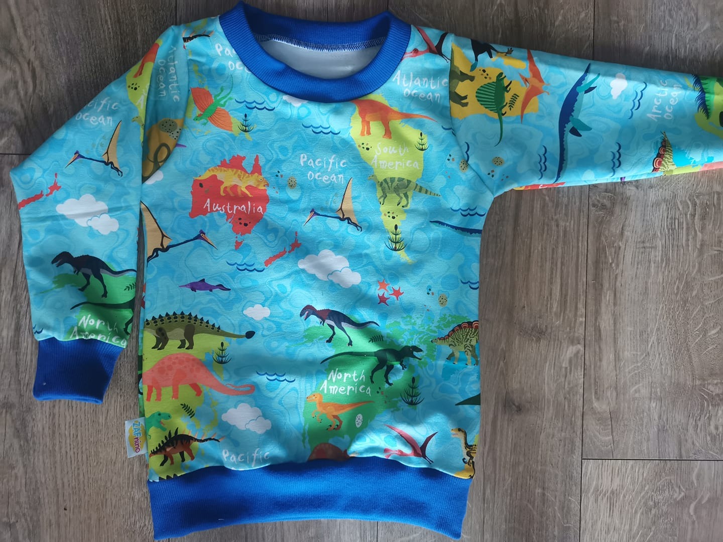 Dziecięca bluza z długim rękawem, z błękitnym tłem i wzorem mapy świata z dinozaurami, leżąca na drewnianej podłodze, widoczna metka ZAFnino na dole.