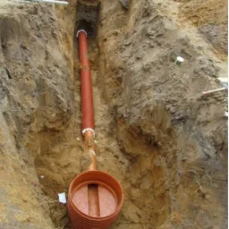 Głęboki wykop z pomarańczową rurą kanalizacyjną i studzienką rewizyjną na dnie, widoczne fragmenty pracowników na górze, ziemia o zróżnicowanej strukturze.