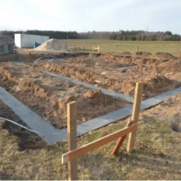 Widok z góry na wykopane rowy pod fundamenty z częściowo wylanym betonem, drewniane szalunki, sterta kostki brukowej i piasku w tle, krajobraz wiejski.