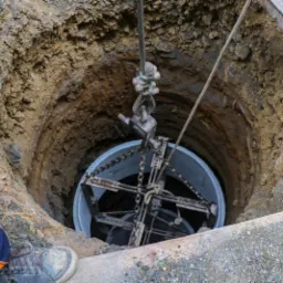 Betonowy krąg studzienny opuszczany do wykopu za pomocą stalowej liny i specjalistycznego uchwytu, widoczne fragmenty odzieży dwóch osób przy krawędzi wykopu.