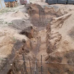 Wykop pod fundamenty z widocznym zbrojeniem, przygotowany do zalania betonem. W tle palety z bloczkami i workami. Widoczne ślady po wykopie w piaszczystej glebie.