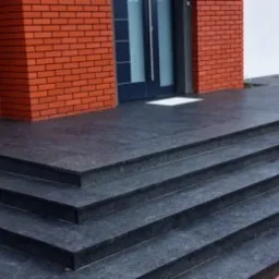 Granitowe schody wejściowe do domu z ceglaną elewacją, wykończone w ciemnoszarym kolorze, z widocznym fragmentem tarasu i zieleni w tle.