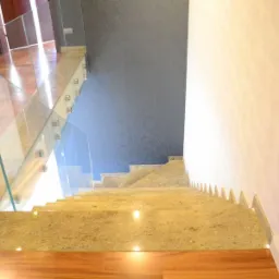 Widok z góry na schody wyłożone polerowanym kamieniem w odcieniach beżu i brązu, zabezpieczone szklaną balustradą mocowaną punktowo, z delikatnym oświetleniem LED w listwach przypodłogowych.