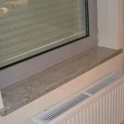 Jasnoszary parapet o drobnej, ziarnistej fakturze, zamontowany pod oknem z żaluzjami, nad grzejnikiem panelowym, widok z boku.