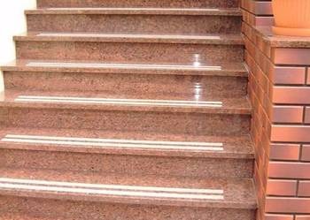 Schody zewnętrzne wyłożone polerowanym granitem w odcieniach brązu i beżu, z antypoślizgowymi paskami, przylegające do ceglanej ściany z donicą na szczycie