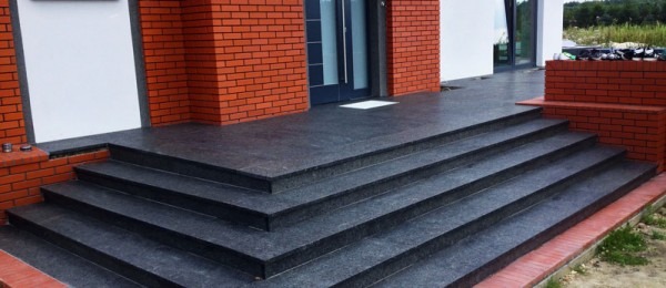 Granitowe schody wejściowe do domu z ceglaną elewacją, wykończone w ciemnoszarym kolorze, z widocznym fragmentem tarasu i zieleni w tle.