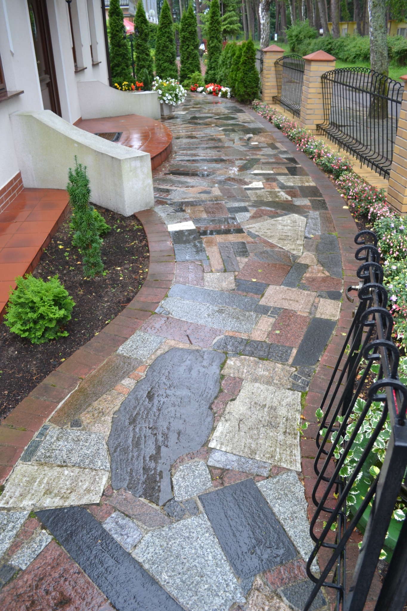 Ścieżka z nieregularnych kamiennych płyt w odcieniach szarości, brązu i czerwieni, obramowana ceglaną krawędzią i roślinami, prowadząca do wejścia domu z zielonymi tujami w tle.