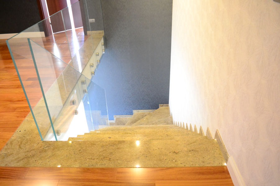 Widok z góry na schody wyłożone polerowanym kamieniem w odcieniach beżu i brązu, zabezpieczone szklaną balustradą mocowaną punktowo, z delikatnym oświetleniem LED w listwach przypodłogowych.