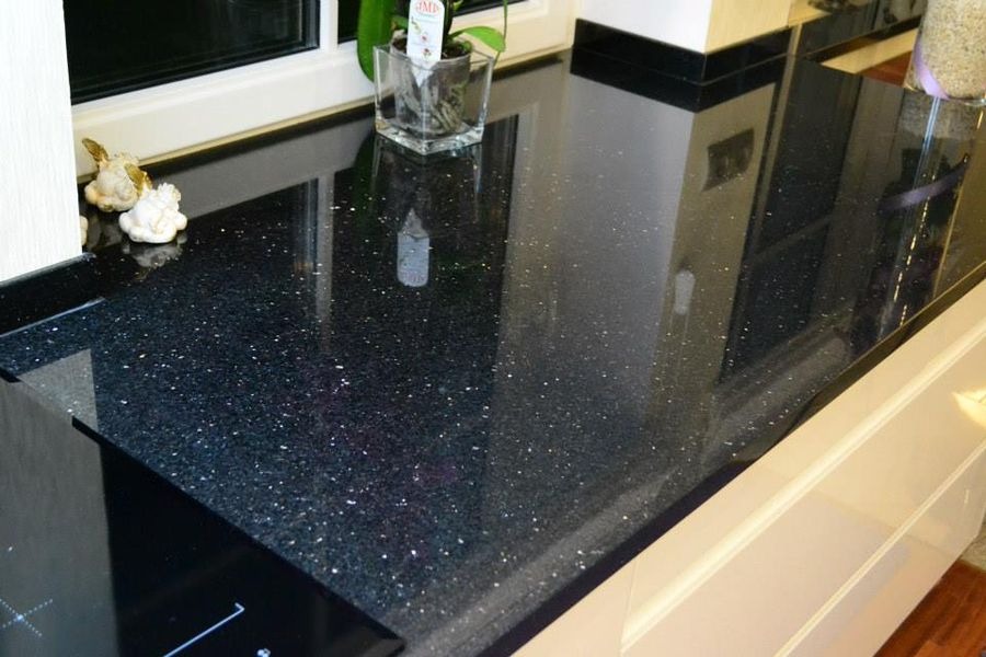 Błyszczący, czarny blat kamienny z drobnymi, srebrnymi inkluzjami, widoczny fragment płyty indukcyjnej, okno z rośliną w tle