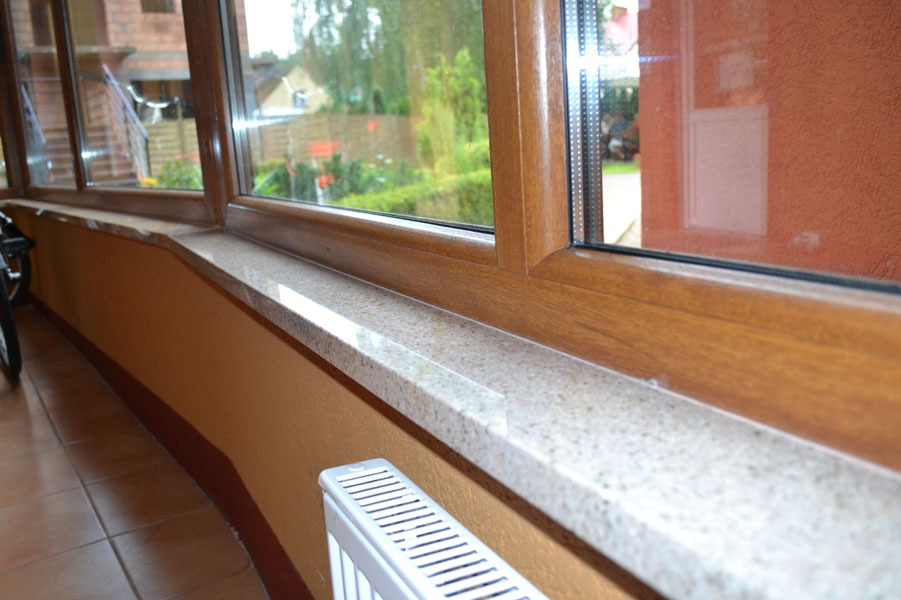 Wnętrze z oknami z brązowymi ramami i jasnym parapetem z nakrapianego materiału, widoczny grzejnik i fragment roweru.