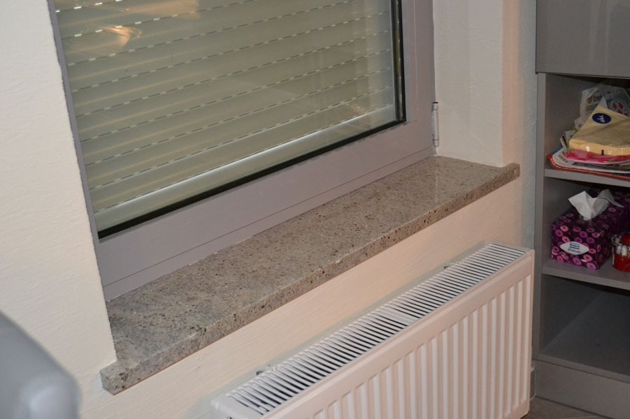 Jasnoszary parapet o drobnej, ziarnistej fakturze, zamontowany pod oknem z żaluzjami, nad grzejnikiem panelowym, widok z boku.