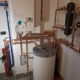 Złożona instalacja grzewcza z kotłem, zasobnikiem ciepłej wody i rurami miedzianymi, widoczne manometry i zawory, zamontowana na ścianie w pomieszczeniu technicznym.