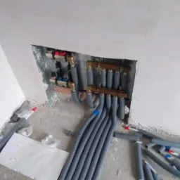 Rozdzielacz ogrzewania podłogowego z szarymi rurami w izolacji, zamontowany w surowym stanie, widoczne przyłącza i elementy regulacyjne.