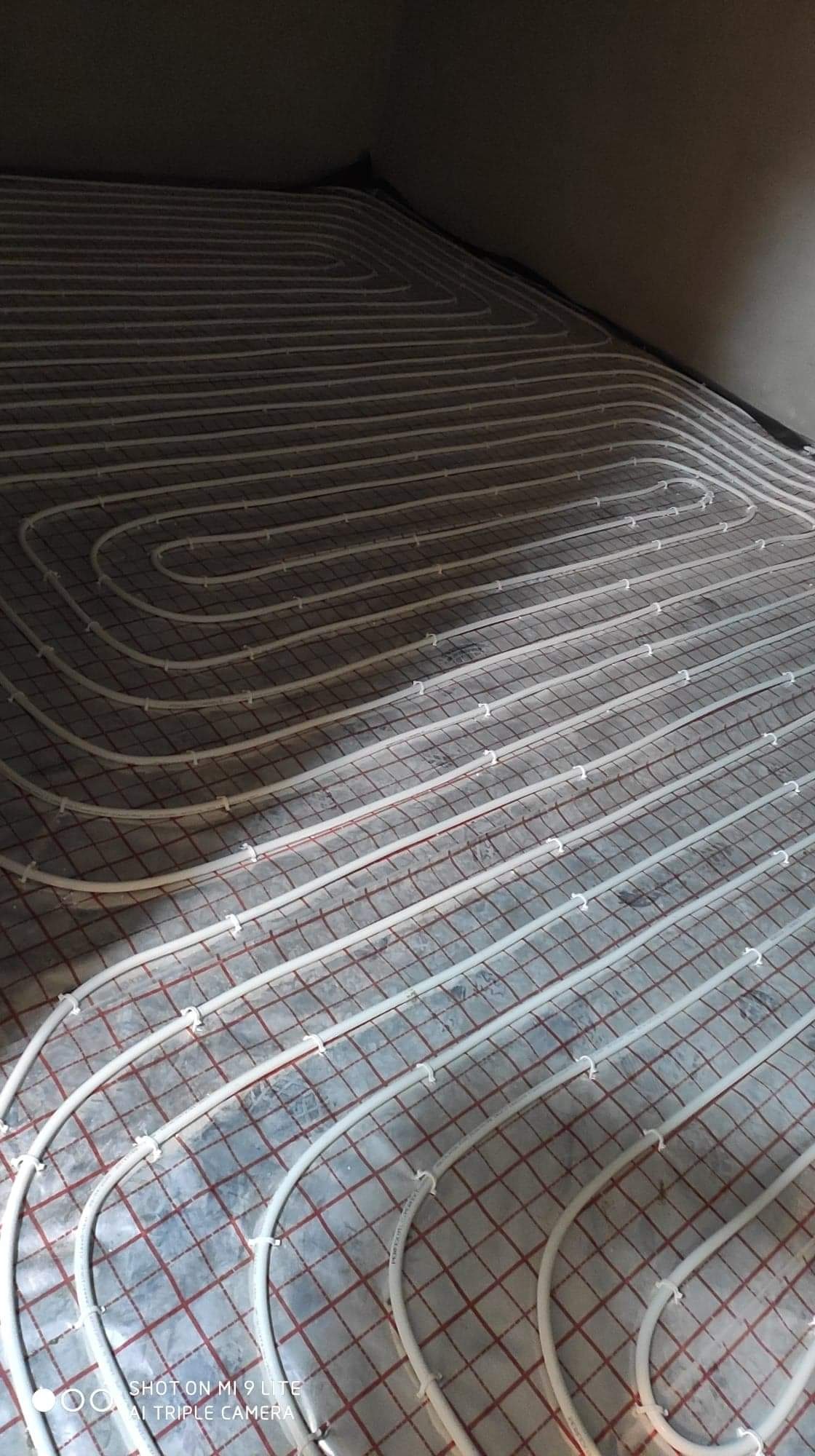 Białe rury ogrzewania podłogowego ułożone w pętle na czerwonej siatce montażowej, widok z góry, surowy stan przed zalaniem wylewką.