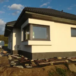 Fragment elewacji domu jednorodzinnego z widocznym oknem narożnym i przygotowanym terenem pod prace wykończeniowe, na tle błękitnego nieba i częściowo widocznego samochodu dostawczego.