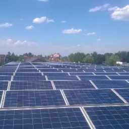 Rozległa instalacja fotowoltaiczna na dachu budynku, widok z góry, błękitne panele pod błękitnym niebem z białymi obłokami, w tle zabudowania mieszkalne i drzewa.