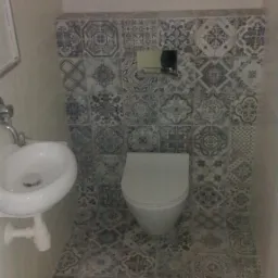 Mała łazienka z umywalką na postumencie i toaletą w zabudowie, wykończona płytkami w stylu patchwork z motywami kwiatowymi i geometrycznymi w odcieniach szarości i błękitu.