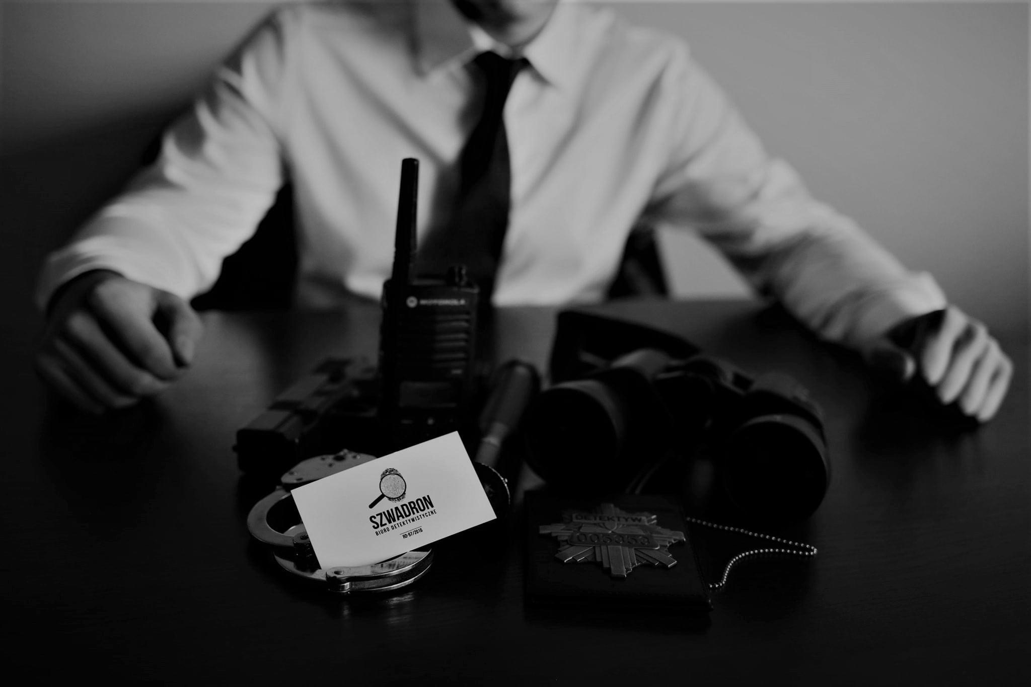 Czarno-białe zdjęcie przedstawia biurko z rekwizytami: pistolet, radiotelefon, lornetka, odznaka detektywa, kajdanki oraz wizytówka z lupą i napisem 'SZWADRON Biuro Detektywistyczne'.