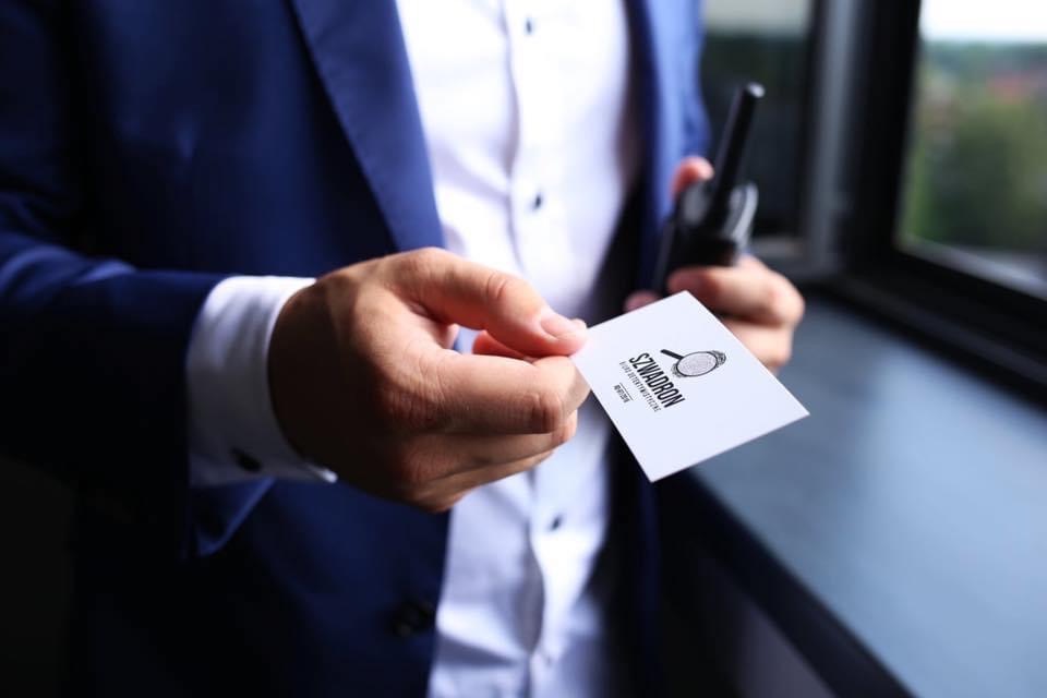 Mężczyzna w granatowym garniturze trzyma wizytówkę z logo firmy detektywistycznej 'Szwadron' i krótkofalówkę.