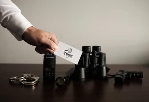 Dłoń w mankiecie białej koszuli trzyma wizytówkę z lupą, obok leżą kajdanki, radiotelefon Motorola, latarka, lornetka i pistolet na ciemnym drewnianym blacie.