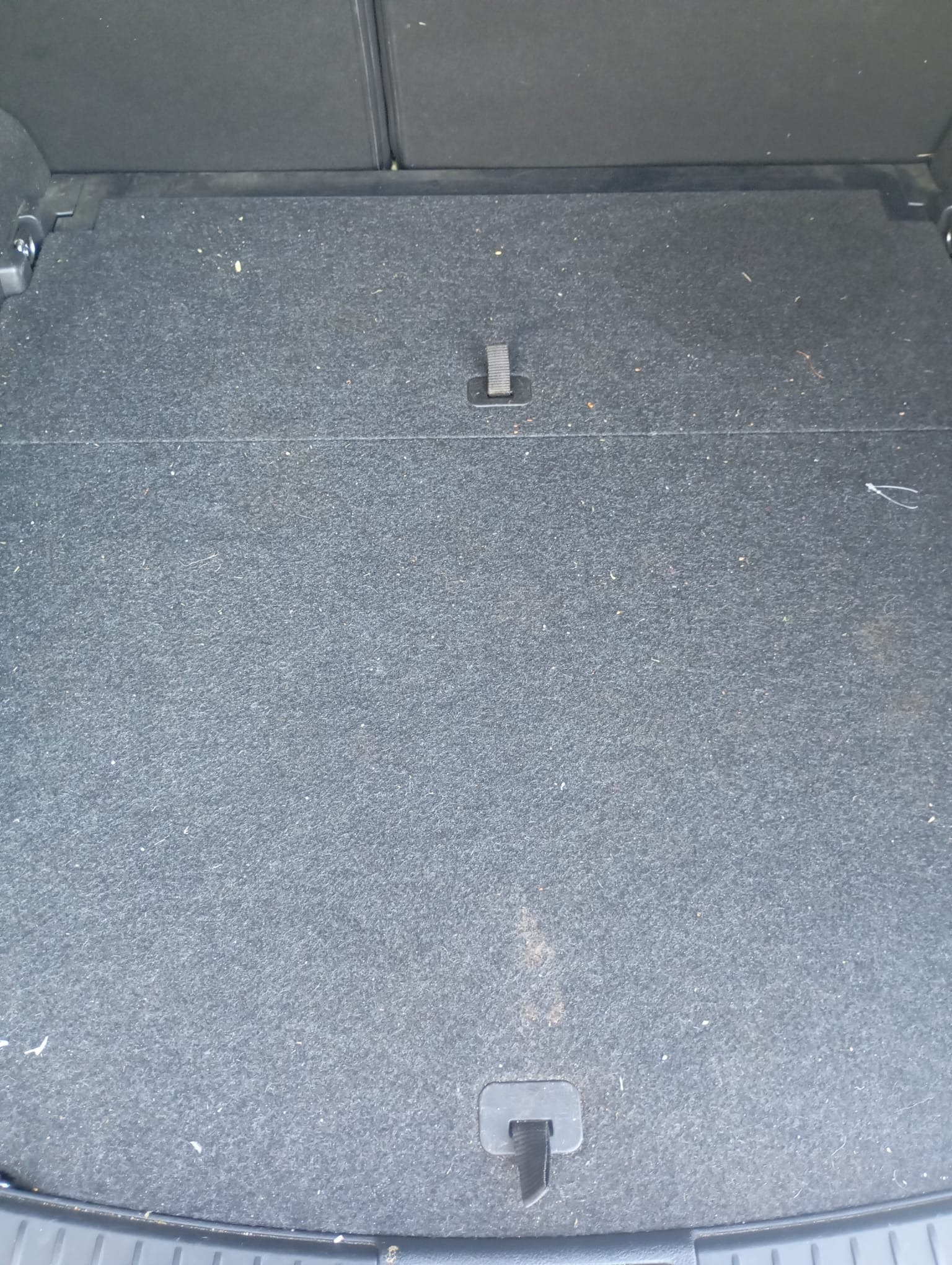 Szara, tekstylna powierzchnia bagażnika samochodowego z widocznymi uchwytami mocującymi i drobnymi zabrudzeniami, ukazująca stan przed czyszczeniem.