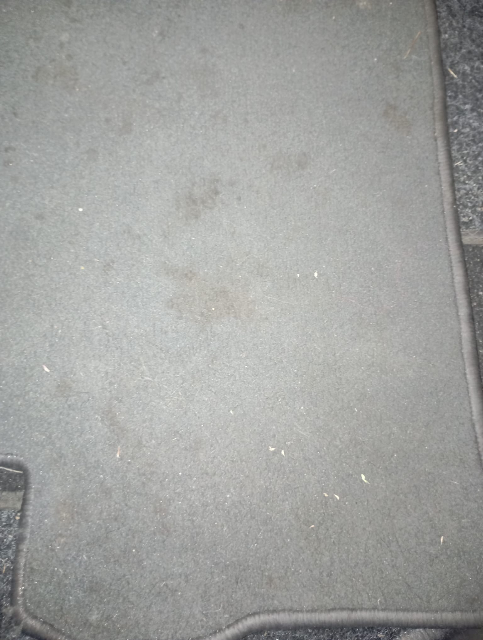 Szary dywanik samochodowy z widocznymi plamami i zabrudzeniami, ukazujący potrzebę odświeżenia i wyprania tapicerki. Widoczne włókna i obrzeże dywanika.