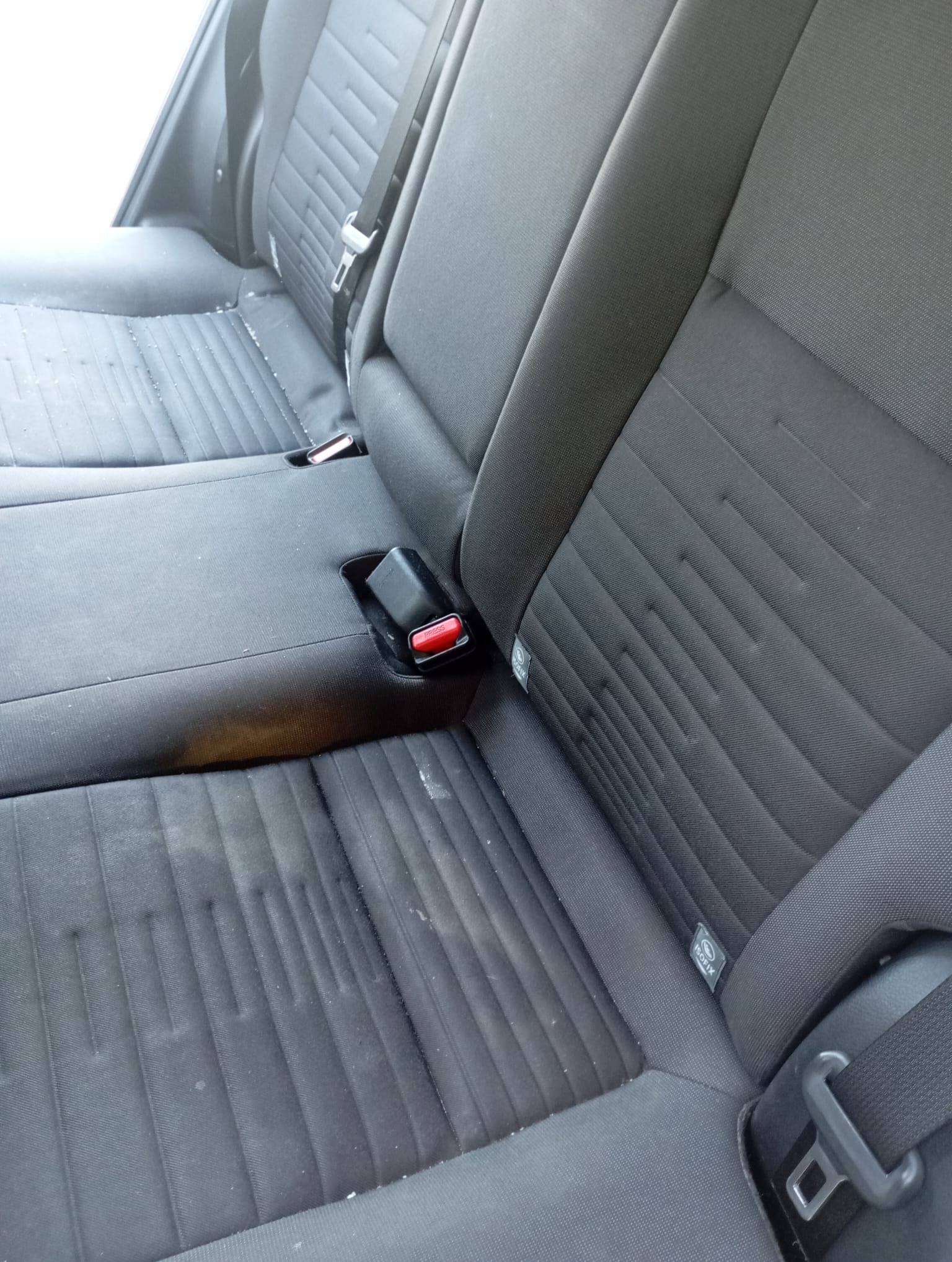 Brudna, tekstylna tapicerka tylnej kanapy samochodu z widocznymi plamami i zabrudzeniami, wymagająca odświeżenia i czyszczenia. Widoczne pasy bezpieczeństwa.