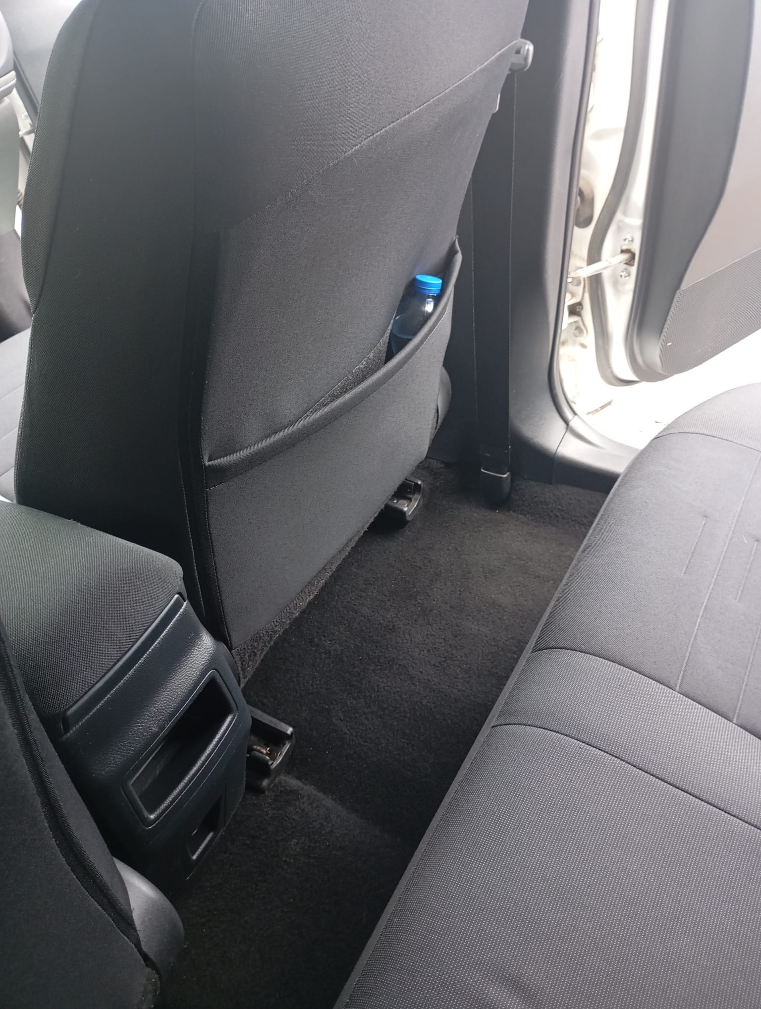 Wnętrze samochodu z czarną tapicerką, widok na tylne siedzenia i podłogę. W kieszeni fotela widoczna butelka wody. Ciemna wykładzina podłogowa.