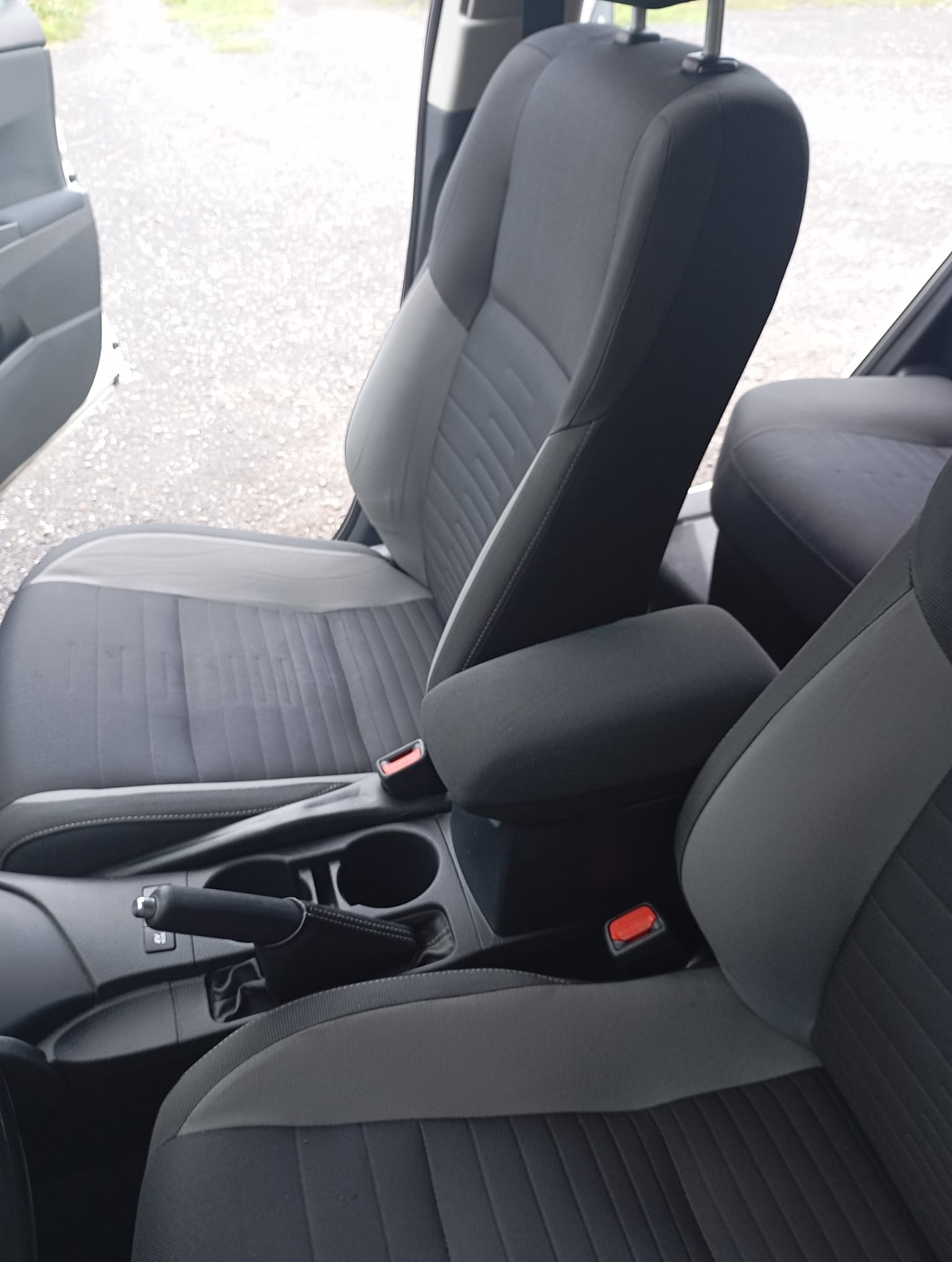 Wnętrze samochodu z szarymi fotelami i podłokietnikiem, widoczne detale tapicerki i konsola środkowa. Otwarta boczna szyba auta.