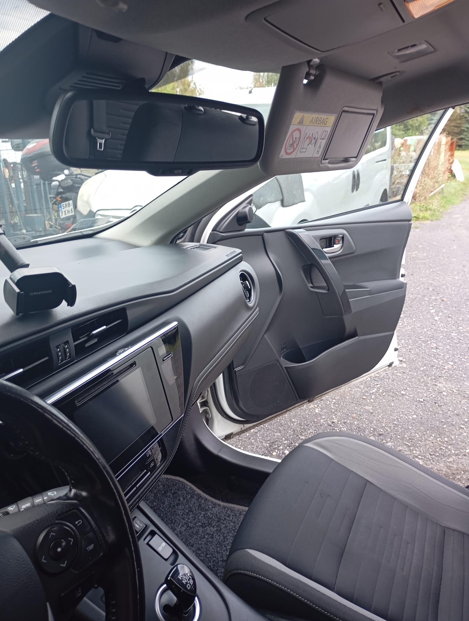 Wnętrze samochodu z otwartymi drzwiami, widoczny kokpit, kierownica, fotel kierowcy z szarą tapicerką i lusterko wsteczne. Na zewnątrz zaparkowany biały van.