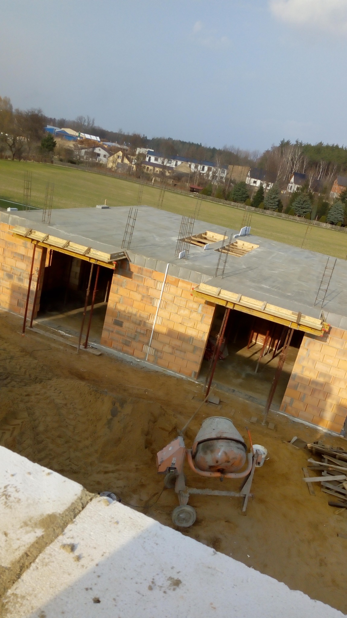 Murowanie ścian z pustaków ceramicznych na budowie domu jednorodzinnego, widoczna betonowa płyta fundamentowa, zbrojenia, podpory szalunkowe i betoniarka na placu budowy.