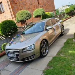 Złoty samochód Audi A8 udekorowany kwiatami i wstążkami, zaparkowany na podjeździe przed domem z cegły, obok zadbanych krzewów.